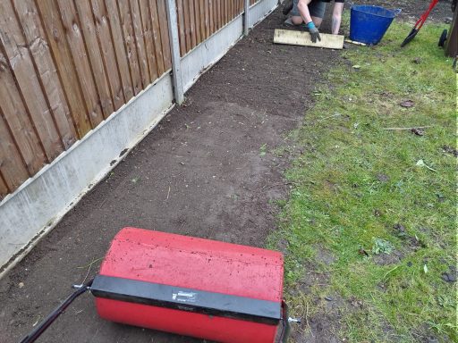 A red roller on freshly prepared soil alongside a wooden fence and tools nearby.